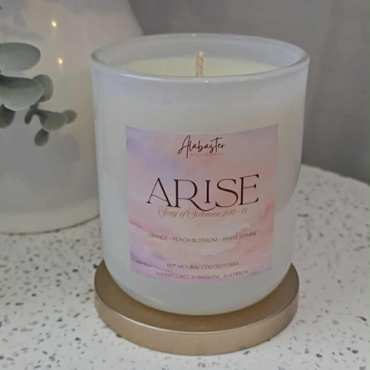 Candle with 'Arise' packaging on a surface with flowers in the background